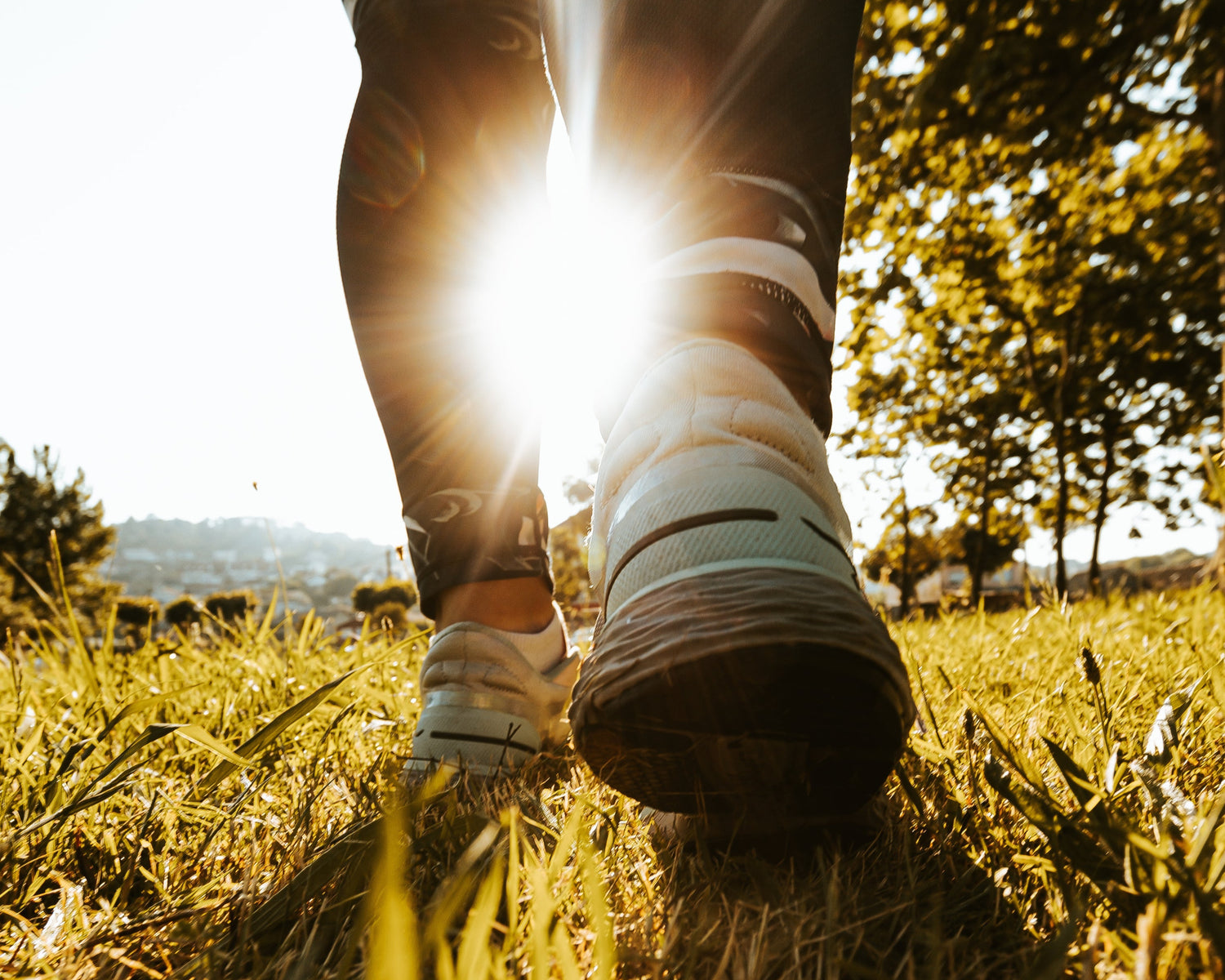 A close up of someone's calves and running shoes on golden grass as the sun peeks between their legs.