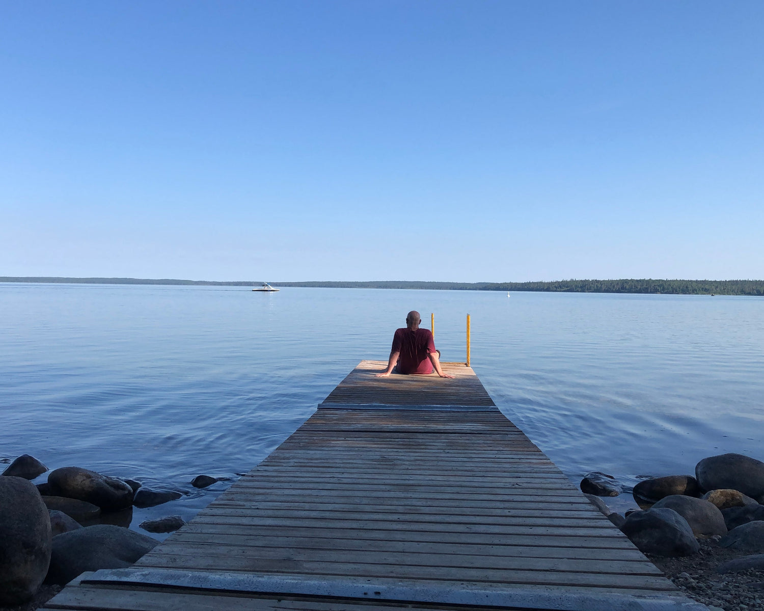 A bald man in a maroon t-shirt sits on the end of a wooden dock overlooking a blue lake and sky. He gazes at a motorboat far off in the distance.