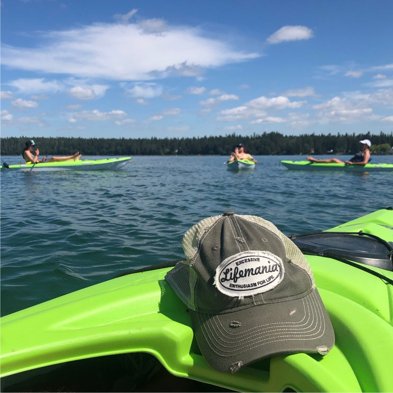 A green and gray trucker hat with a 'Lifamanics' logo sits on the bright green bow of a kayak in the foreground, with a lake, kayakers, and forest in the background.