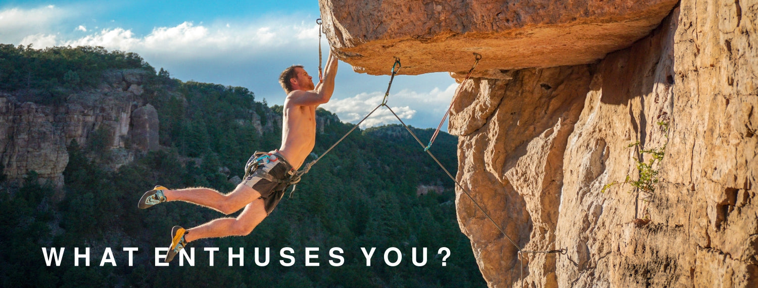 A shirtless man climbing or hanging from a rock face outdoors, with canyon mountains and blue sky in the background. Large white text at the bottom reads, 'WHAT ENTHUSIASMS YOU?'.
