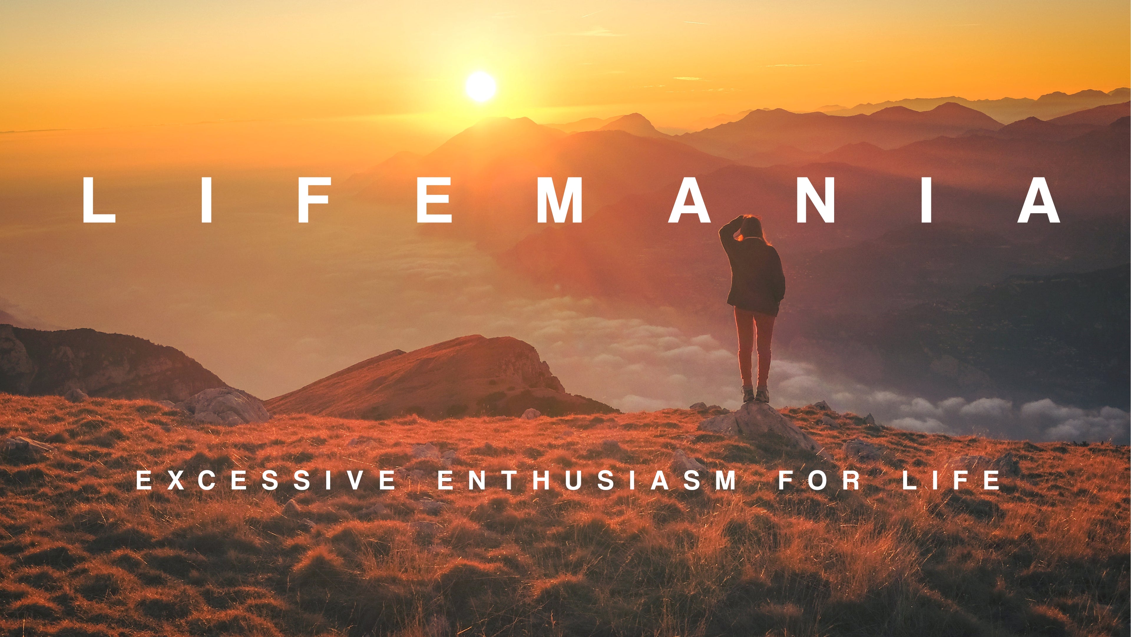 A woman stands on a small boulder atop a mountain, looking past the cloud tops of a deep valley and taking in the sun setting behind the distant mountaintops.
