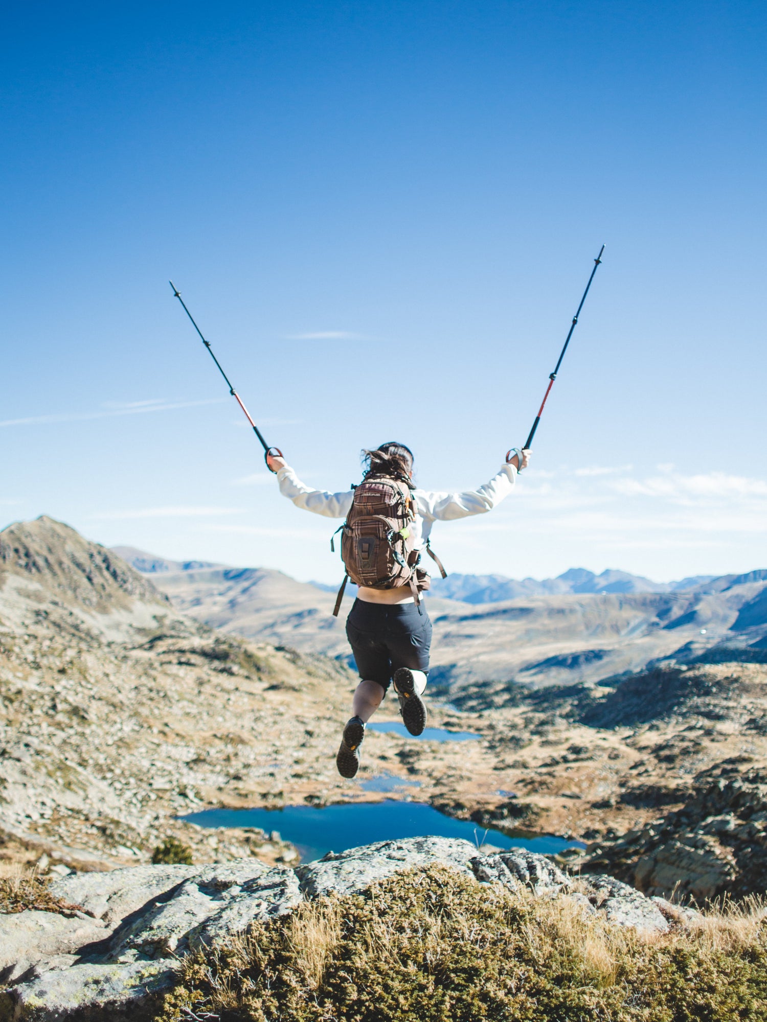 A female backpacked hiker, atop a rocky hilltop, jumped up and is suspended in the air with her hiking poles pointing to the pure blue sky at the end of her outstretched arms. A small lake is below in the valley surrounded by small rolling coulees.